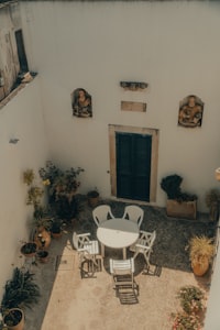 A sunlit courtyard with white chairs, a round table, potted plants and green shutters in Puglia, southern Italy.